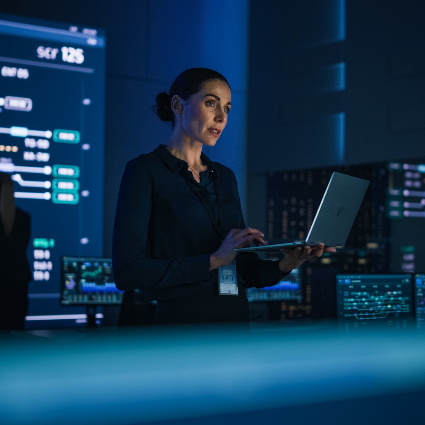 Beautiful Confident Female Project Manager works on Laptop Standing in Telecommunications System Control Room. In Background Big Screen Showing Infographics of Infrastructure, Charts, System Analysis