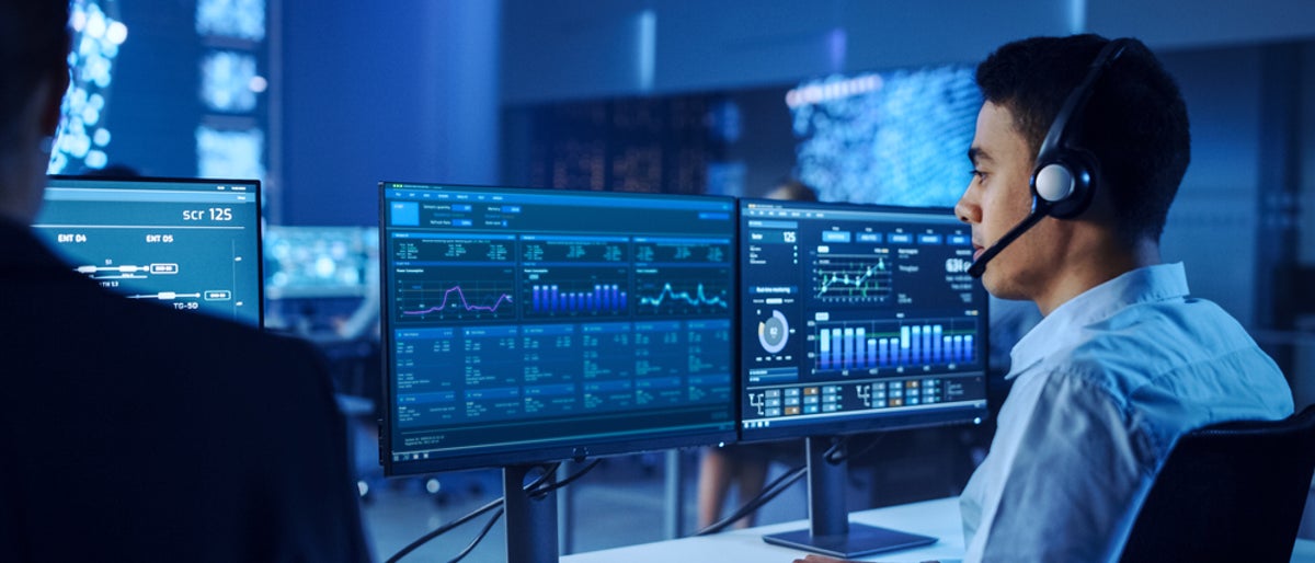 Confident Male Data Scientist Works on Personal Computer Wearing a Headset in Big Infrastructure Control and Monitoring Room. Young Engineer in a Business Call Center Office Room with Colleagues.