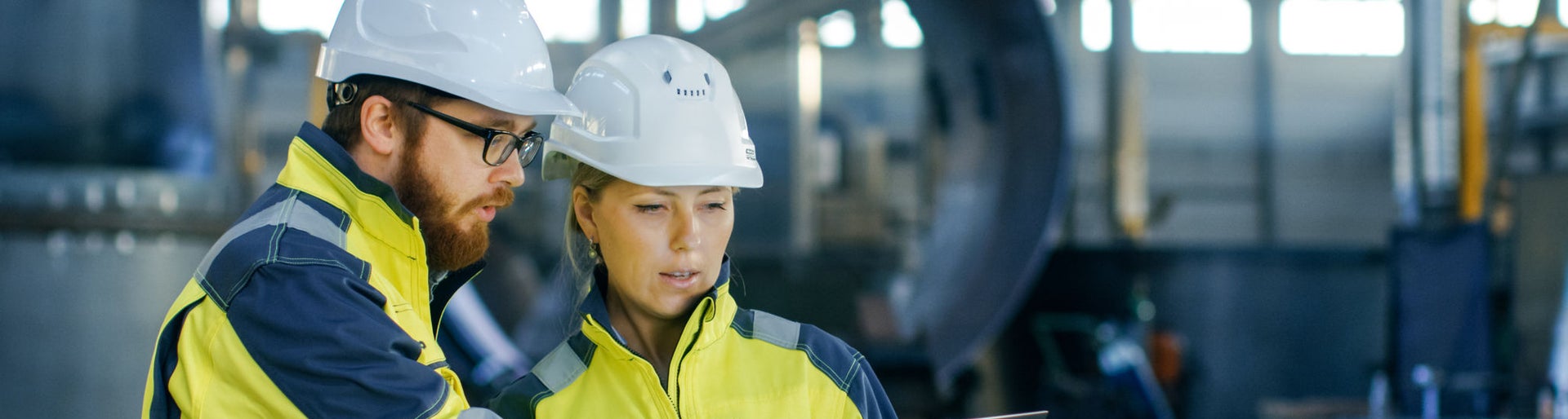 Male and Female Industrial Engineers in Hard Hats Discuss New Project while Using Laptop. They Make Showing Gestures.They Work in a Heavy Industry Manufacturing Factory.