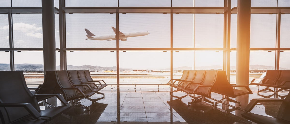 Wide-angle view of a modern aircraft gaining the altitude outside the glass window facade of a contemporary waiting hall with multiple rows of seats and reflections indoors of an airport terminal