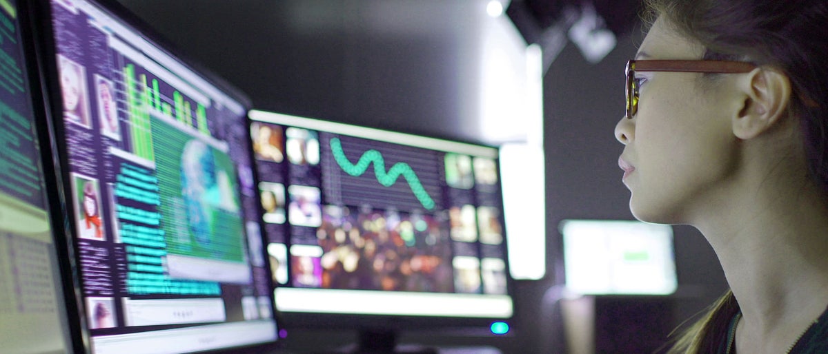 Close up stock image of a young asian woman sitting down at her desk where she’s surrounded by 3 large computer monitors displaying out of focus images of people as thumbnails; crowds; graphs & scrolling text.