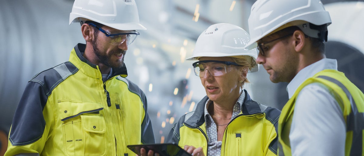 Three Heavy Industry Engineers Stand in Pipe Manufacturing Factory, Use Digital Tablet Computer, Have Discussion. Large Pipe Assembled. Design and Construction of Oil, Gas and Fuels Transport Pipeline