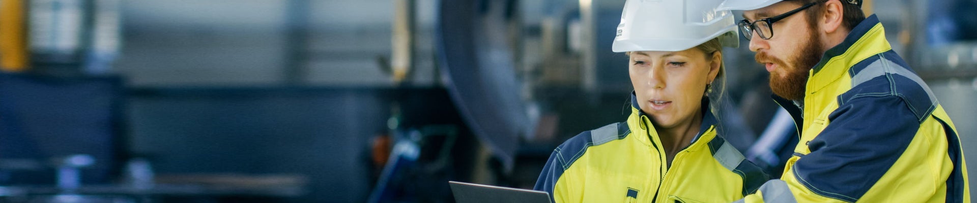 Male and Female Industrial Engineers in Hard Hats Discuss New Project while Using Laptop. They Make Showing Gestures.They Work in a Heavy Industry Manufacturing Factory.
