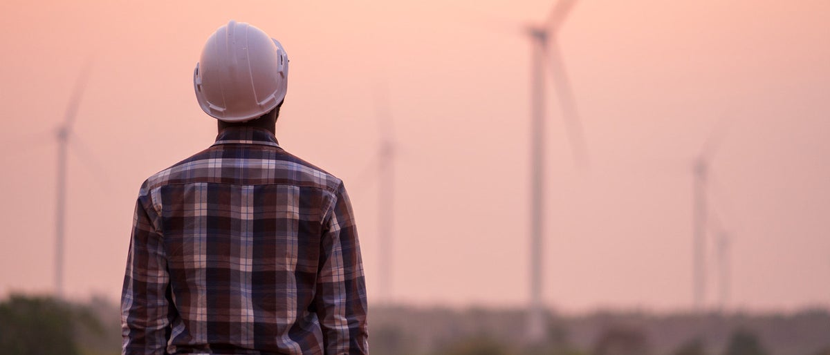African engineer wearing white hard hat standing with digital tablet against wind turbine on sunny day