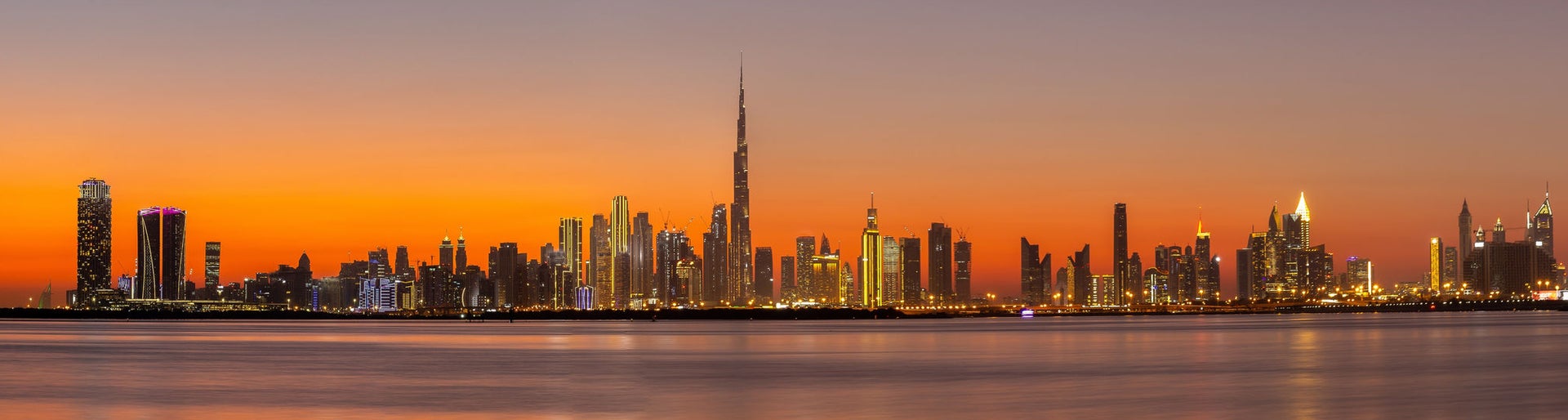 Panorama of Dubai Business Bay skyline at night after sunset with colorful illuminated buildings and calm Dubai Creek water.