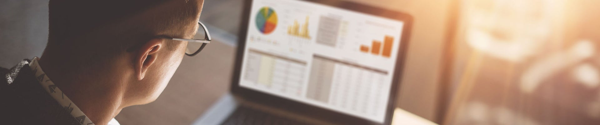 Young finance market analyst in eyeglasses working at sunny office on laptop while sitting at wooden table.Businessman analyze document in his hands.Graphs and diagramm on notebook screen.Blurred